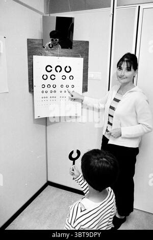 Esame della scuola medica del test dell'occhio, Whizgle News, Dutch Desk, Paesi Bassi, 1950 - 2000 il 06-1999. Questi argomenti vengono visualizzati nell'immagine. In uno spazio clinico luminoso, una donna si trova accanto a un ampio grafico pieno di varie forme circolari in bianco e nero, progettate per testare la visione. Ha un comportamento coinvolgente, vestito con un cardigan di colore chiaro e un top a righe, che trasuda calore e avvicinabilità. I suoi capelli sono modellati in modo ordinato, e lei sta indicando la cartella, probabilmente spiegando qualcosa a un bambino di fronte a lei. Il bambino, che sembra curioso e leggermente concentrato, ha un piccolo strumento che assomiglia a Foto Stock