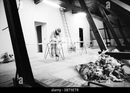 Ex ristrutturazione. Municipio B'wijk Beverwijk Nederland, Whizgle News, Dutch Desk, Paesi Bassi, 1950 - 2000 il 13-12-1990. L'immagine include questi argomenti. In una spaziosa mansarda parzialmente rifinita, c'è un'atmosfera di operosità e creatività. La camera presenta travi in legno che aggiungono un fascino rustico, contrastando con gli elementi moderni sparsi ovunque. Al centro, una figura è piegata su un lungo tavolo, meticolosamente concentrata sul loro compito. Vestito con abiti da lavoro casual, questo individuo è circondato da strumenti e materiali, indicativi di un progetto in corso. La tabella è clu Foto Stock