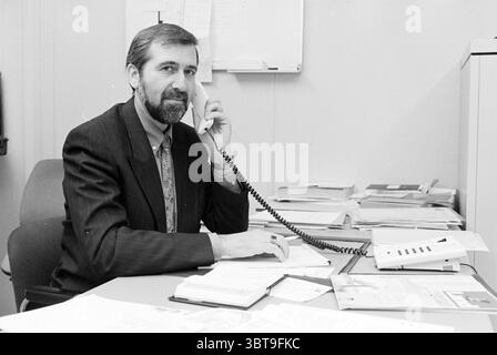 Uomo sul posto di lavoro dietro la scrivania, Whizgle News, Dutch Desk, Paesi Bassi, 1950 - 2000 il 20-12-1994. L'immagine mostra questi argomenti. In un ambiente d'ufficio modesto, un uomo siede su una scrivania disordinata, impegnato in una conversazione telefonica. Ha una barba ben curata ed è vestito con un abito scuro, accentuato da una cravatta a motivi geometrici. La sua espressione focalizzata suggerisce che è profondamente impegnato nel pensiero o nel discutere di una questione importante. La scrivania è piena di pile di documenti, alcuni dei quali sono ordinatamente organizzati, mentre altri sembrano disgustati. Alcuni file rimangono aperti, a indicare che il lavoro è in corso. Alla sua destra, un piccolo blocco note Foto Stock