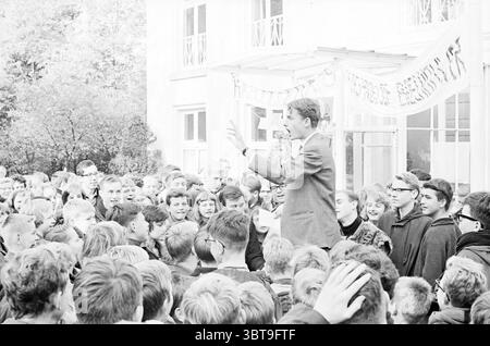 Elezioni del Consiglio scolastico Mendelcollege e Karel van Manderlyceum Giochi giovanili e giovanili scuole, Whizgle News, Dutch Desk, Paesi Bassi, 1950 - 2000 il 11-10-1963. Questi sono gli argomenti nell'immagine. La scena cattura un vivace raduno all'aperto, dove un uomo si trova in primo piano, coinvolgendo un pubblico affascinato. È vestito con una tuta intelligente, gestendo animatamente con una mano alzata, suggerendo di essere a metà discussione o radunando la folla. La sua espressione è animata, riflette passione o entusiasmo. Il pubblico è composto principalmente da giovani, molti dei quali si appoggiano, Foto Stock