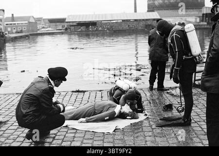 Esami in immersioni vigili del fuoco vigili del fuoco, Whizgle News, Dutch Desk, Paesi Bassi, 1950 - 2000 il 18-11-1971. Questi sono gli argomenti nell'immagine. In un ambiente oscuro e industriale, un gruppo di individui viene raccolto vicino a un corpo d'acqua, avvolto in una spessa atmosfera ricoperta che proietta una luce grigia e silenziosa sulla scena. L'acqua riflette il cielo opaco, creando uno stato d'animo malinconico. In prima linea, una figura giace su una coperta adagiata sui ciottoli, vestita con un lungo indumento di colore chiaro. C'è un senso di urgenza in quanto due persone si prendono cura di loro, una è eseguire ciò che appare Foto Stock