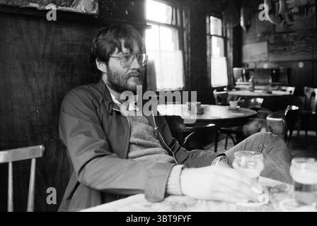Tour attraverso NH - Hoorn Enkhuizen Medemblik Enkhuizen, Whizgle News, Dutch Desk, Paesi Bassi, 1950 - 2000 al 20-06-1981. Questi sono gli argomenti nell'immagine. La scena è ambientata in un ambiente rustico e poco illuminato di un pub o caffetteria che trasuda un ambiente caldo, invitante ma leggermente nostalgico. La luce soffusa si riversa dalle grandi finestre, gettando ombre delicate sulle pareti di legno scuro e sui vari mobili sparsi in tutto lo spazio. In primo piano, un uomo si siede casualmente, appoggiandosi su una sedia di legno. Ha un'espressione premurosa, indossando grandi occhiali incorniciati che accentuano il suo conte Foto Stock