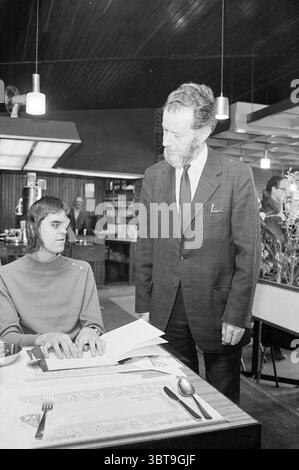 Qualcuno che legge Braille al ristorante Beverwijk Station Beverwijk Paesi Bassi., Whizgle News, Dutch Desk, Paesi Bassi, 1950 - 2000. Questi sono gli elementi dell'immagine. La scena presenta un ristorante con un'estetica contemporanea della metà del secolo. Un giovane uomo siede a un tavolo, le sue mani poggiano su un menu. Indossa un pullover moderno e minimalista che completa la sua espressione premurosa. L'acconciatura del giovane uomo è indicativa dell'epoca, con frange più lunghe. Accanto a lui c'è un uomo più anziano, vestito bruscamente con una tuta a quadri, che trasuda autorità o forse curiosità. La o Foto Stock