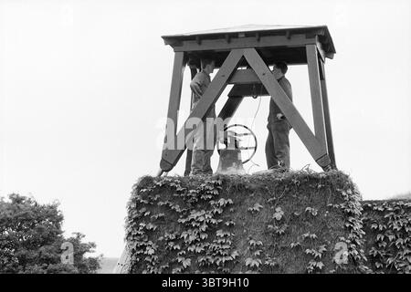 Appeso la campana al cimitero Onorario Bloemendaal, Whizgle News, Dutch Desk, Paesi Bassi, 1950 - 2000 il 19-06-1991. L'immagine include questi argomenti. La scena cattura un ambiente rustico all'aperto caratterizzato da un campanile costruito con travi in legno, che forma una struttura robusta che sostiene un tetto inclinato. La torre sorge in cima a un lussureggiante tumulo, ricoperto di vibrante edera verde che ricopre la struttura in natura. Due figure sono posizionate vicino al campanello, apparendo impegnate in una conversazione o magari collaborando a qualche compito. Sono vestite con toni tenui, che si fondono armoniosamente con il Foto Stock