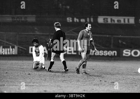 AJAX - Juventus 1-1 Football Ajax, Whizgle News, Dutch Desk, Paesi Bassi, 1950 - 2000 il 01-03-1978. L'immagine mostra questi argomenti. La scena cattura un momento vivido su un campo da calcio sotto le luci artificiali. Il passo è contrassegnato da linee bianche luminose, in contrasto con la superficie scura e bagnata. In primo piano, un giocatore si inginocchia a terra, la sua postura suggerisce un momento di frustrazione o stanchezza. È vestito con una maglia di colore chiaro abbinata a pantaloncini scuri, mentre il secondo giocatore si trova nelle vicinanze, indossando una maglia più scura a contrasto. L'arbitro, posizionato tra di loro, è rivestito Foto Stock