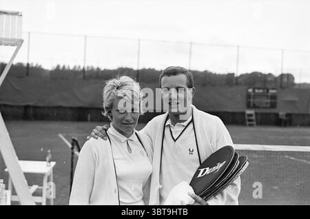 Tennis Brederode III Tennis, Whizgle News, Dutch Desk, Paesi Bassi, 1950 - 2000 il 07-1965. Questi sono gli elementi dell'immagine. In un vivace ambiente esterno, due persone si trovano fianco a fianco su un campo da tennis ben tenuto. Lo sfondo rivela un confine a rete e una distesa erbosa, con un accenno di elementi esterni sfocati che suggeriscono un ampio paesaggio oltre. La prima persona, una donna dai capelli biondi corti e elastici, indossa una polo bianca e un cardigan leggero coordinato drappeggiato casualmente sulle spalle. La sua espressione è amichevole, con un leggero sorriso che trasmette calore Foto Stock