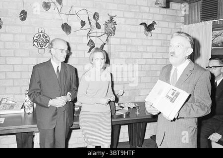 Si rivolge a una coppia e si congratula., Whizgle News, Dutch Desk, Paesi Bassi, 1950 - 2000. Questi sono gli elementi dell'immagine. In un ambiente interno accogliente, tre persone sono presenti in modo prominente in una conversazione. Lo sfondo è costituito da pareti in mattoni caldi e testurizzati che aggiungono un senso di casa alla scena. Un tavolo in legno adornato con piccoli oggetti in vetro si erge in primo piano, suggerendo un incontro sociale. A sinistra, un uomo con gli occhiali, vestito con un abito scuro, sembra ascoltare con attenzione. Ha un'espressione premurosa, contribuendo a creare un'atmosfera serena. Accanto a lui, a Foto Stock