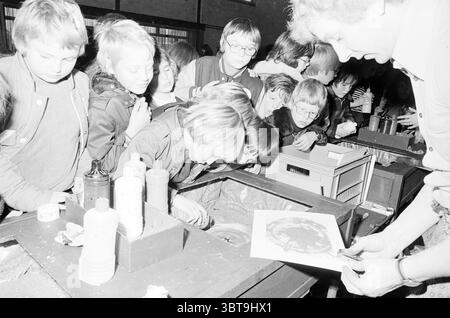 Gruppo di bambini in escursione., Whizgle News, Dutch Desk, Paesi Bassi, 1950 - 2000. L'immagine include questi argomenti. La scena cattura un vivace raduno di bambini, che sono raggruppati da vicino, i loro volti un mix di curiosità ed eccitazione. Il gruppo è prevalentemente maschile, con alcune ragazze intervallate tra loro. I loro capelli variano da corti e ordinati a più lunghi e duri, mostrando una gamma di stili tipici dell'epoca. In primo piano, una donna è impegnata con i bambini, tenendo un pezzo di carta circolare che sembra raffigurare una sorta di disegno o modello. Lei si inclina sl Foto Stock