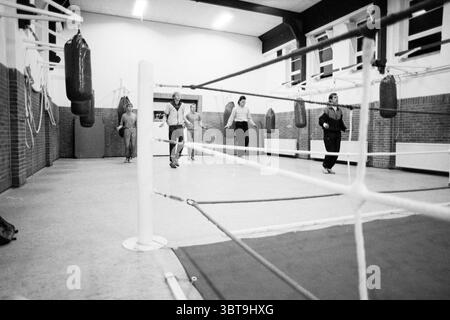Boxing club de Ring Eksterstraat IJmuiden Boxing IJmuiden Nederland, Whizgle News, Dutch Desk, Paesi Bassi, 1950 - 2000 il 14-03-1989. L'immagine include questi argomenti. La scena cattura una palestra di boxe con un forte senso di movimento e attività. In primo piano, un anello di boxe bianco è contrassegnato da corde che si estendono lungo la scena, conducendo l'occhio verso due individui che camminano lungo il bordo dell'anello. Le loro posture sono rilassate e allo stesso tempo decise, suggerendo una conversazione informale o un cameratismo. Lo sfondo è caratterizzato da diversi sacchetti di punzonatura pesanti sospesi dal c Foto Stock