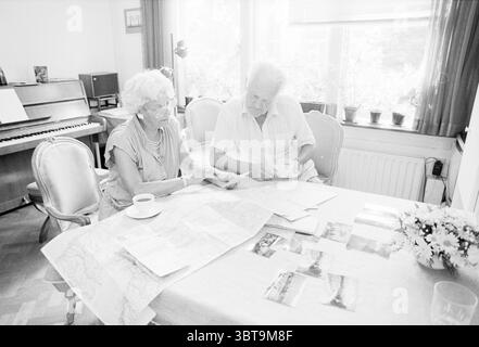 Famiglia Brown fine delle vacanze Bloemendaal Holiday Vacidaymakers Bloemendaal, Whizgle News, Dutch Desk, Paesi Bassi, 1950 - 2000 il 06-07-1989. Questi argomenti vengono visualizzati nell'immagine. In una stanza soleggiata, due persone anziane sono sedute a un tavolo adornato con mappe e fotografie. La tovaglia, un morbido colore crema, contrasta con la ricca struttura in legno della tavola sottostante. Sono impegnati nella conversazione, le loro espressioni riflettono un senso di nostalgia e contemplazione. La donna, con i capelli e gli occhiali bianchi ricci, ha un sorriso caldo mentre indica una particolare sezione della mappa che diffonde bef Foto Stock