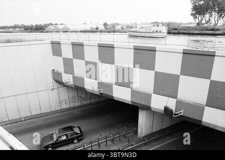 Acquedotto Leimuiderdijk. Costruzione di strade messa in strada Leimuiderdijk, Whizgle News, Dutch Desk, Paesi Bassi, 1950 - 2000 il 09-08-1989. L'immagine contiene questi argomenti. La scena presenta un'ampia vista di un paesaggio urbano strutturato, dominato principalmente da una grande struttura in cemento multistrato che funge da ingresso o uscita sotterraneo. La superficie in alto presenta un suggestivo motivo composto da quadrati alternati in sfumature di grigio, che contrasta con gli elementi lisci in cemento. Sotto questa facciata a motivi geometrici, un singolo veicolo scuro viene catturato in movimento, allontanandosi dal Foto Stock