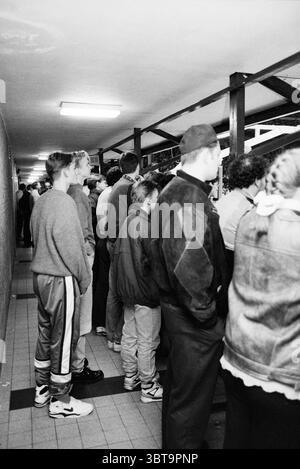 Public HBW [Haarlem Basketball Week], Whizgle News, Dutch Desk, Paesi Bassi, 1950 - 2000 il 04-01-1992. L'immagine mostra questi argomenti. La scena è ambientata in uno stretto corridoio, caratterizzato da un soffitto basso e da un'architettura semplice e funzionale. Le pareti sono composte da una superficie silenziosa e leggermente ruvida, che conferisce un'atmosfera industriale allo spazio. Le luci fluorescenti proiettano un bagliore luminoso e uniforme, creando un forte contrasto con gli abiti più scuri della folla. Un gruppo eterogeneo di persone si trova vicino, di fronte a un tavolo o a un bancone alla fine del corridoio. L'accordo forma un ti Foto Stock