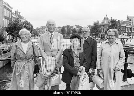 Crociera per anziani con storie di vacanza e foto di gruppo crociera visite turistiche voli turistici, Whizgle News, Dutch Desk, Paesi Bassi, 1950 - 2000 il 19-07-1984. L'immagine contiene questi argomenti. La scena cattura un piccolo gruppo di cinque persone che si trovano insieme su un lungomare, circondato da un pittoresco paesaggio urbano. La gente, vestita con un mix di cappotti chiari e neutri, mostra un'aria di cameratismo. Le loro espressioni vanno dal contentamento ai sorrisi aperti, suggerendo un momento condiviso di gioia o nostalgia. Lo sfondo presenta edifici caratteristici e affascinanti Foto Stock