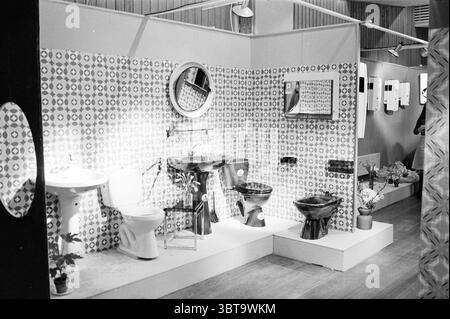 Mostra di primavera degli interni de Linge Interior, Whizgle News, Dutch Desk, Paesi Bassi, 1950 - 2000 su 27-03-1975. Questi argomenti vengono visualizzati nell'immagine. La scena presenta un display da bagno progettato per mostrare vari tipi di impianti e design. Le pareti sono adornate da un'audace piastrella a motivi geometrici che presenta forme geometriche in colori contrastanti, evocando un senso di fascino retrò. A sinistra, accanto ad una piccola pianta decorativa in vaso, si trova un classico WC bianco che aggiunge un tocco di verde all'ambiente. Accanto ad esso, sono esposti un elegante lavandino nero e un lavandino nero coordinato, la loro superficie lucida Foto Stock
