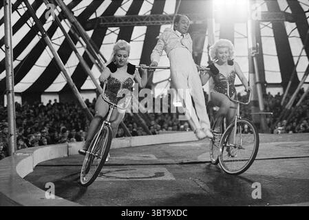 Circus Boltini Circus, Whizgle News, Dutch Desk, Paesi Bassi, 1950 - 2000 il 20-04-1969. Questi sono gli argomenti nell'immagine. In un ambiente da circo vivace, l'atmosfera è viva di energia ed eccitazione. Due artisti, vestiti con costumi scintillanti che brillano sotto le luci della tenda, guidano le biciclette fianco a fianco. I loro abiti sono decorati con paillettes e glitter, riflettendo una gamma di colori come argento e rosa, creando un effetto abbagliante. Al centro, un uomo vestito con una tuta bianca immacolata esegue un trucco impressionante, apparentemente sfidando la gravità mentre salta tra i due ciclisti. Suo Foto Stock
