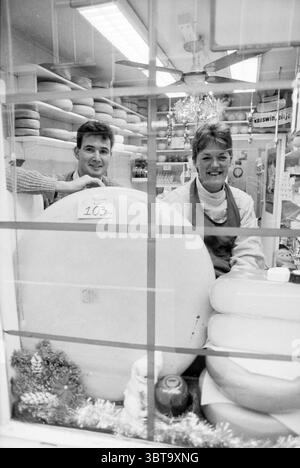 Negozio di formaggi con 103 kg di formaggio, Whizgle News, Dutch Desk, Paesi Bassi, 1950 - 2000 il 12-12-1990. L'immagine mostra questi argomenti. La scena cattura un caratteristico interno di un negozio, caratterizzato da un'atmosfera calda e invitante. Dietro una grande finestra, due persone si trovano accanto a un'impressionante esposizione di forme di formaggio. La prima persona, un uomo con i capelli scuri e un sorriso sottile, si appoggia su una grande forma di formaggio, apparendo rilassato e amichevole. Accanto a lui, una donna dai capelli di colore chiaro, legata con cura, si erge con fiducia, indossando un grembiule che suggerisce il suo ruolo nel negozio. Lo schema di colori Foto Stock