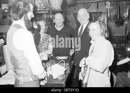 First Beaujolais in the Good Hour Wine, Whizgle News, Dutch Desk, Paesi Bassi, 1950 - 2000 il 15-11-1975. L'immagine include questi argomenti. La scena si sviluppa in uno spazio intimo e poco illuminato, probabilmente un bar rustico o un punto di ritrovo, caratterizzato da travi in legno e arredi vintage che emana un calore invitante. L'atmosfera sembra vivace, infuso con l'energia del cameratismo. Al centro, un gruppo di cinque persone si impegna in una conversazione animata. A sinistra, un uomo con una barba prominente è vestito con una camicia bianca e pantaloni scuri, la sua postura aperta e invitante mentre interagisce con gli altri. Foto Stock