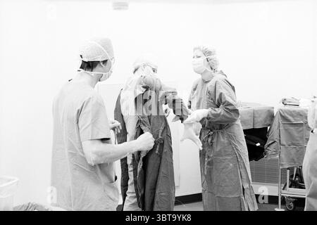 Operazione Johannes de Deo, Whizgle News, Dutch Desk, Paesi Bassi, 1950 - 2000 il 03-05-1990. Questi argomenti vengono visualizzati nell'immagine. In un ambiente clinico, un gruppo di medici professionisti è impegnato nella preparazione di una procedura chirurgica. L'ambiente è caratterizzato da pareti rigide e sterili che riflettono un'illuminazione bianca e luminosa, creando un'atmosfera pulita e clinica. Due persone indossano scrub chirurgici abbinati, che appaiono in sfumature di blu, mentre una terza persona è rivestita in un abito chirurgico verde. Ogni professionista ha i propri capelli coperti, con cappucci chirurgici e maschere facciali in posizione, e. Foto Stock