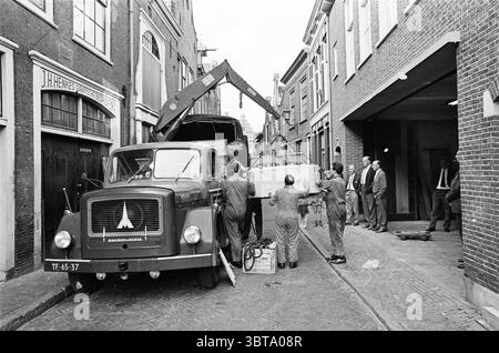 Posizionamento della nuova sezione della pressa. Haarlems Dagblad ecc. e IJmuider Courant e Koerier. Facade J.H. Henkes Graphic Companies, Whizgle News, Dutch Desk, Paesi Bassi, 1950 - 2000 il 24-07-1963. L'immagine mostra questi argomenti. In una strada stretta e trafficata fiancheggiata da edifici in mattoni, un camion d'epoca si erge in posizione prominente, il cui retro è sollevato da una gru meccanica. Il camion, decorato con un logo distinto, aggiunge un tocco di nostalgia alla scena. Intorno al veicolo vengono raccolte diverse persone, impegnate in quello che sembra essere uno sforzo collaborativo per scaricare un oggetto di grandi dimensioni. Il popolo, vestito a metà dei 20t Foto Stock