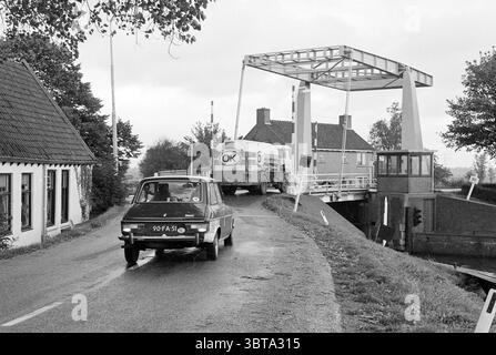 Autovettura e autocisterna su una strada stretta vicino al ponte degli impianti di risalita di Nauerna Nauerna, Whizgle News, Dutch Desk, Paesi Bassi, 1950 - 2000 nel 1978. L'immagine include questi argomenti. In un ambiente tranquillo ma leggermente coperto, una strada stretta si snoda attraverso una tranquilla zona rurale. Da un lato, un'affascinante casa rustica con un ripido tetto spiovente e tegole scure, le sue pareti dipinte in una tenue tonalità di bianco. Il verde lussureggiante circonda la casa, con l'erba che ondeggia dolcemente nella brezza dolce, che aggiunge un'atmosfera tranquilla. Una piccola auto d'epoca, caratterizzata dalla sua forma compatta e dalle caratteristiche arrotondate, guida al meglio Foto Stock