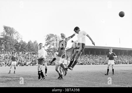 Royal HFC - Sports Club Emma Football, Whizgle News, Dutch Desk, Paesi Bassi, 1950 - 2000 il 25-06-1961. L'immagine mostra questi argomenti. La scena cattura un momento dinamico durante una partita di calcio, ambientata in un grande campo aperto. Diversi giocatori sono in azione, mostrando una serie di posture atletiche. Al centro, un giocatore alto con una maglia bianca salta in testa alla palla, mostrando un mix di determinazione e concentrazione. I suoi compagni di squadra, vestiti con maglie scure, sono posizionati strategicamente intorno al campo, le loro espressioni sono tese e impegnate nello svolgimento del gioco. In primo piano, un portiere in contropartita Foto Stock
