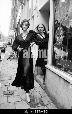 Exest. Boutiques Amsterdam Exterior, Whizgle News, Dutch Desk, Paesi Bassi, 1950 - 2000 il 04-12-1973. Questi argomenti sono mostrati nell'immagine. La scena cattura una strada stretta e animata, con marciapiedi acciottolati che brillano sotto una luce soffusa e diffusa, che accenna a una giornata ricoperta. Su un lato, un vetrino in vetro presenta un elegante manichino vestito da un elegante abito nero, con maniche suggestive e una posa suggestiva che attira l'attenzione. Le caratteristiche del manichino sono nitide ed espressive, impreziosite da accessori che ne esaltano il look audace. In piedi appena fuori dal negozio, un Wo Foto Stock