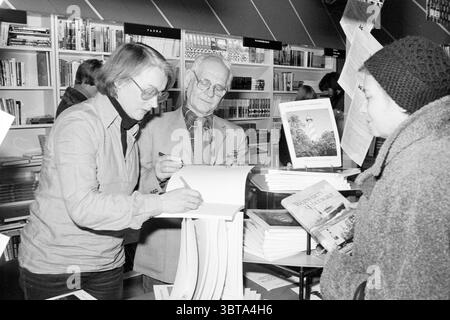 Libro per la firma 'Dutch Lighthouse' in IJmuiden, Whizgle News, Dutch Desk, Paesi Bassi, 1950 - 2000 il 16-02-1979. L'immagine include questi argomenti. In una vivace libreria, si apre una scena piena di interazioni interessanti e dettagli degni di nota. Da un lato, una donna siede ad un tavolo rotondo circondato da pile di libri, la sua espressione si concentra come scrive sulle pagine di un libro che si trova di fronte a lei. Indossa una camicia button-up a maniche lunghe di colore chiaro che contrasta con i toni più profondi dell'ambiente circostante. I suoi capelli sono modellati in modo semplice, evidenziando il suo sguardo intenzionale. Trasversale Foto Stock