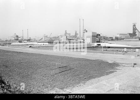 Nuova strada con stazione di pompaggio sulle macchine e le fabbriche del canale IJmuiden, Whizgle News, Dutch Desk, Paesi Bassi, 1950 - 2000 al 20-02-1975. Questi argomenti sono mostrati nell'immagine. La scena presenta un vasto panorama industriale caratterizzato da grandi strutture e macchinari. Tra gli elementi di spicco vi sono torreggianti fumogeni e lunghi edifici angolari che dominano lo skyline, a indicare un impianto di produzione o lavorazione. L'architettura appare utilitaria, con grandi superfici piatte e decorazioni minimali, enfatizzando la funzionalità rispetto all'estetica. La tavolozza dei colori è Foto Stock