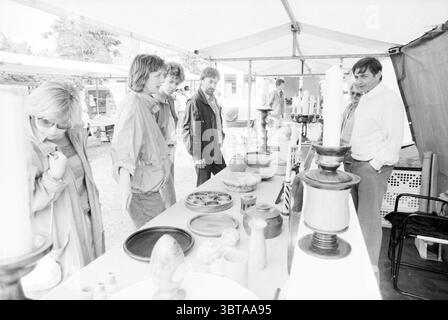 Art Market Spaarndam Markt Spaarndam, Whizgle News, Dutch Desk, Paesi Bassi, 1950 - 2000 il 18-07-1987. Questi sono gli argomenti nell'immagine. Sotto una tenda bianca, si apre una scena di mercato, piena di una varietà di ceramiche fatte a mano e oggetti di artigianato disposti sui tavoli. La superficie dei tavoli mostra una serie di pezzi in ceramica, ciotole, vasi e sculture artistiche, ognuno realizzato con vetrate uniche che riflettono i toni naturali della terra come i marroni profondi, i gialli tenui e i verdi tenui. Un piccolo gruppo di persone, in piedi intorno ai tavoli, sembra impegnato in una conversazione. Sono casual nell'abbigliamento Foto Stock