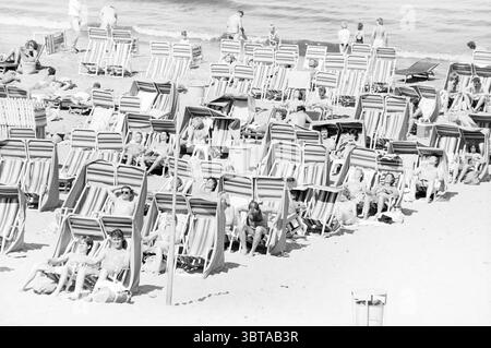 Spiaggia Zandvoort Spiaggia e spiagge Zandvoort, Whizgle News, Dutch Desk, Paesi Bassi, 1950 - 2000 il 04-07-1987. L'immagine contiene questi argomenti. La scena raffigura un vivace ambiente da spiaggia, pieno di una serie di sedie a sdraio allineate in modo ordinato su calde spiagge sabbiose. Le sedie, adornate con tessuti a righe luminose in colori contrastanti, sono occupate da varie persone che si rilassano sotto il sole. Alcune sono metà reclinate, crogiolate alla luce del sole, mentre altre sono sedute in posizione eretta, impegnate in una conversazione o semplicemente godendovi la vista sull'oceano. Sullo sfondo, onde dolci si infrangono contro la riva, crea Foto Stock