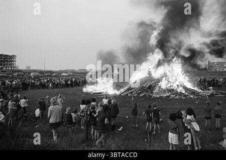 Capanne bruciate nel campo vacanze Schalkwijk giochi vacanze giorni, Whizgle News, Dutch Desk, Paesi Bassi, 1950 - 2000 il 09-08-1968. Questi sono gli elementi dell'immagine. Un grande falò domina il primo piano, torreggiando con fiamme e fumo folto e vorticoso che si libra verso l'alto nel cielo. Il fuoco è costruito con pallet e casse di legno, creando uno spettacolo caotico ma affascinante. Intorno alle fiamme, un gruppo eterogeneo di persone si erge in un semicerchio, i cui volti sono illuminati dal caldo bagliore del fuoco. La folla è un mix di adulti e bambini, con una varietà di età A. Foto Stock