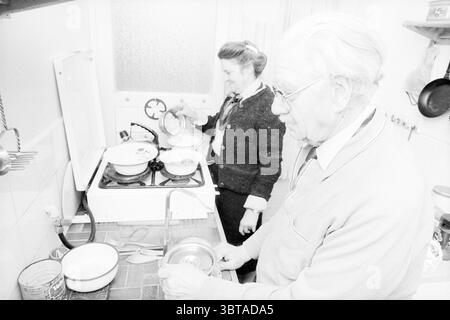 Il signor Selders riceve il cibo consegnato a casa sua da Mrs. Faster Food and Eateries, Whizgle News, Dutch Desk, Paesi Bassi, 1950 - 2000 il 18-02-1987. Questi sono gli elementi dell'immagine. In un ambiente accogliente, due persone sono impegnate nella preparazione dei pasti. La scena viene catturata prevalentemente in tonalità di bianco e nero, conferendole una qualità nostalgica e senza tempo. A sinistra, una donna sta davanti alla stufa, i suoi capelli sono stati modellati con cura. Indossa un cardigan su una camicia con colletto, trasudando un senso di calore e di domesticità. Si concentra intensamente su una pentola, magari controllando il contenuto o aggiungendo un dentro Foto Stock
