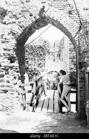 Ruin of Brederode Visitors Ruin, Whizgle News, Dutch Desk, Paesi Bassi, 1950 - 2000 il 15-07-1969. Questi argomenti vengono visualizzati nell'immagine. La scena è ambientata in un ambiente rustico e storico, caratterizzato da pareti in pietra sbriciolate che formano un arco, rivelando i resti di una vecchia struttura. Le pietre mostrano varie sfumature di grigio e marrone, alcune intemperie e coperte di muschio, suggerendo età e decadimento. La luce del sole si riversa attraverso l'arco, proiettando ombre attenuate sul terreno. In primo piano, un ponte di legno attraversa un'apertura stretta, le sue tavole disposte parallelamente, che conduce al cuore della S Foto Stock
