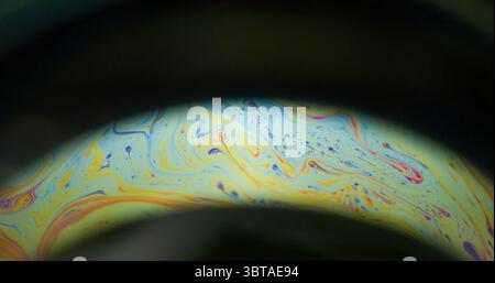 Sapone iridescente. Arco arcobaleno. La lente rotonda colorata dipinge il flusso caotico del colorante misto su sfondo nero. Luce astratta sfocatura notturna giallo-blu. Foto Stock