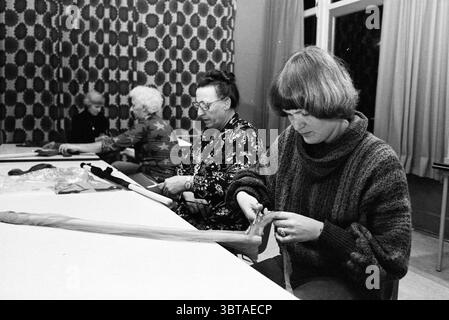 Neighborhood Evening de Lichtboei H'kerk quartieri centri comunitari negozi locali centri di formazione, Whizgle News, Dutch Desk, Paesi Bassi, 1950 - 2000 on 26-10-1978. L'immagine include questi argomenti. In un ambiente interno con luce soffusa, diverse persone sono impegnate in un'attività artigianale intorno a un lungo tavolo. La tavolozza di colori predominante presenta tonalità calde e motivi ricchi, con uno sfondo che mostra un tessuto adornato da intricati disegni geometrici in colori contrastanti. Tre donne siedono a tavola, ognuna assorbita dai propri compiti. La donna all'estrema destra, vestita con una maglia grossa Foto Stock