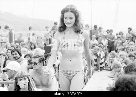 Beach inn sfilata di moda e Miss Elections concorso di bellezza Zo Voort sfilata di moda Zandvoort, Whizgle News, Dutch Desk, Paesi Bassi, 1950 - 2000 il 28-07-1979. Questi argomenti sono mostrati nell'immagine. La scena cattura una vivace atmosfera da spiaggia piena di una folla diversificata. Al centro della composizione c'è una giovane donna che si affida con sicurezza su una pista improvvisata, indossando un elegante bikini Vichy che accentua la sua figura. I suoi lunghi capelli cadono in onde morbide, incorniciando il suo viso, con un pizzico di determinazione giocosa. Intorno a lei c'è un mucchio di spettatori, le loro espressioni una miscela di adm Foto Stock