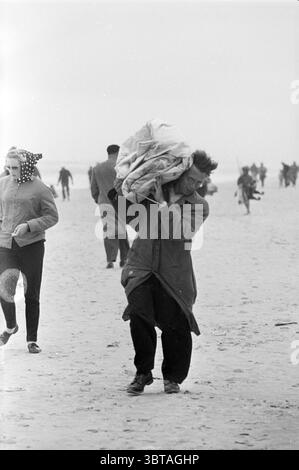 Fishermen on the Beach Beach and Beaches Fisheries and other fishmongers fishing, Whizgle News, Dutch Desk, Paesi Bassi, 1950 - 2000 il 14-01-1963. Questi sono gli argomenti nell'immagine. In questa scena costiera, i toni morbidi e silenziosi del grigio e del bianco dominano l'atmosfera, creando un senso di freschezza e luce soffusa. La spiaggia si estende, sabbiosa e piatta, con dolci onde che si infrangono sullo sfondo. Un uomo è al centro della scena, strisciando intenzionalmente sulla sabbia. Porta un grande oggetto impacchettato sulla sua spalla, stretto in tessuto, suggerendo un'aria di determinazione e di sforzo. H Foto Stock