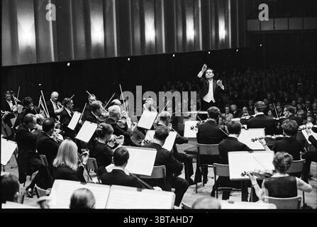 Direttore ospite della North Philharmonic Orchestra Music, Whizgle News, Dutch Desk, Paesi Bassi, 1950 - 2000 il 24-11-1967. Questi argomenti sono mostrati nell'immagine. La scena cattura un momento in una sala da concerto piena di energia e attesa. In primo piano, un conduttore si trova con sicurezza nella parte anteriore, bracci sollevati come se guidasse l'insieme con fervore. È vestito con un classico smoking nero, trasuda un'aria di autorità e passione. Dietro di lui, un ensemble orchestrale è disposto in formazione semicerchio, il loro abbigliamento formale scuro che si fonde armoniosamente con l'atmosfera sofisticata del V Foto Stock