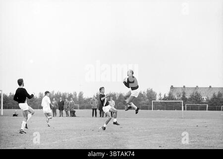 OG - Bloemendaal Voetbal VV Schoten Onze Gezellen Renova, Whizgle News, Dutch Desk, Paesi Bassi, 1950 - 2000 il 21-09-1969. L'immagine contiene questi argomenti. In un vasto campo aperto che sembra essere un campo sportivo erboso, un gruppo di individui è impegnato in un'intensa partita di calcio. La scena è dominata dalle varie sfumature di verde dell'erba, intervallate dal bianco intenso delle uniformi dei giocatori. Due giocatori sono in primo piano: Uno, vestito in bianco e nero, salta in aria, mostrando atletismo mentre tenta di prendere un colpo. La sua espressione è concentrata Foto Stock