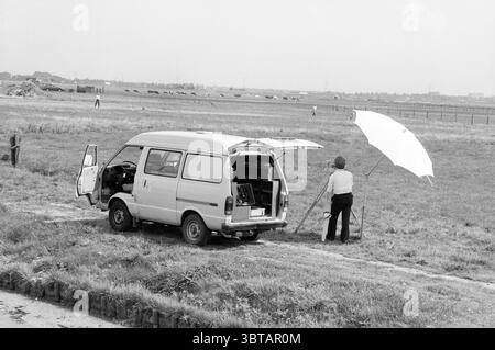 Segui le indicazioni per Velsertunnel lungo la A9. Indicazioni stradali costruzione di strade piantagione stradale Beverwijk Rijksweg A22 Paesi Bassi, Whizgle News, Dutch Desk, Paesi Bassi, 1950 - 2000 on 24-08-1987. L'immagine include questi argomenti. La scena mostra un paesaggio tranquillo e aperto sotto un cielo luminoso e parzialmente nuvoloso. Un furgone di colore chiaro, leggermente intemprato, è parcheggiato a lato di un sentiero sterrato che conduce alla distesa erbosa. La porta posteriore del furgone è aperta, rivelando un interno modesto, accennando ad alcune attrezzature o rifornimenti all'interno. Nelle vicinanze, una persona si trova accanto al furgone, tenendo Foto Stock