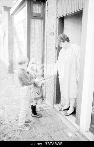 Vendita di porte da bambini IJmuiden bambini case per bambini festa per bambini vendita per bambini IJmuiden Paesi Bassi, Whizgle News, Dutch Desk, Paesi Bassi, 1950 - 2000 il 27-09-1984. L'immagine mostra questi argomenti. La scena cattura un momento fuori da una porta, incorniciata da un modesto edificio in mattoni. A sinistra, la luce del sole si riversa attraverso una grande finestra con tende a motivi geometrici, creando un caldo bagliore che contrasta con le tonalità fredde dell'ambiente circostante. Il pavimento è lastricato di pietre quadrate, indicativo di un quartiere vivace. In primo piano, due bambini stanno vicini. T Foto Stock