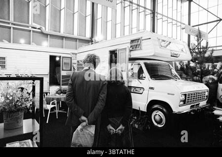Caravan - Rai Amsterdam Camping, Whizgle News, Dutch Desk, Paesi Bassi, 1950 - 2000 il 12-12-1978. Questi sono gli elementi dell'immagine. La scena cattura uno spazio espositivo al coperto, pieno di attività. L'attenzione è rivolta a un camper bianco d'epoca, ben esposto, che trasuda un senso di nostalgia. La sua superficie liscia e lucida riflette le luci circostanti, donandole un'invitante luce soffusa. Di fronte al camper, una coppia sta stretta l'una con l'altra, le spalle si voltano verso lo spettatore. L'uomo, vestito con un abito a righe scure, appare rilassato, con le mani casualmente nascoste dietro di lui. T Foto Stock