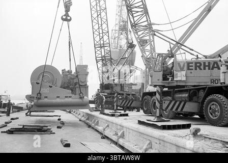 Argano di carico sulla barca IJmuiden carico e scarico IJmuiden Paesi Bassi, Whizgle News, Dutch Desk, Paesi Bassi, 1950 - 2000 il 20-06-1979. Questi argomenti sono mostrati nell'immagine. La scena cattura un vivace lungomare industriale, avvolto in un'atmosfera silenziosa a causa del cielo coperto. La tavolozza dei colori è prevalentemente grigia, enfatizzando gli elementi in acciaio e cemento dell'ambiente. Le gru torreggianti dominano la composizione, con le loro lunghe braccia meccaniche che raggiungono il cielo, sagomate sullo sfondo nuvoloso. In primo piano, un grande carico circolare viene sollevato da uno dei Foto Stock