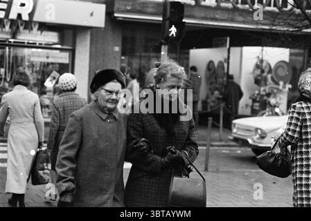 Shopping Public., Whizgle News, Dutch Desk, Paesi Bassi, 1950 - 2000. Questi argomenti vengono visualizzati nell'immagine. In una vivace scena di strada, due donne più anziane camminano a stretto contatto, apparendo coinvolte in una conversazione. Il loro abbigliamento è composto da cappotti scuri e testurizzati, entrambi indossano cappelli eleganti che riflettono un'eleganza classica. Una donna ha un colletto rifinito in pelliccia che aggiunge un tocco di eleganza al suo outfit. Intorno a loro si trova un vivace ambiente urbano pieno di attività. Lo sfondo presenta una vetrina con un luminoso display, con una serie di prodotti che attirano l'attenzione. Peopl Foto Stock
