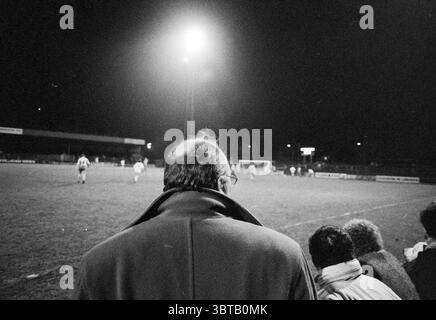 Telstar - Veendam Football Telstar, Whizgle News, Dutch Desk, Paesi Bassi, 1950 - 2000 il 10-03-1990. Questi sono gli argomenti nell'immagine. Sulla scena, una folla di spettatori si riunisce in un luogo sportivo all'aperto durante la notte. L'attenzione è rivolta a un uomo in primo piano, visto da dietro, che indossa un lungo cappotto scuro. La sua testa calva riflette la luce brillante emessa da un proiettore sospeso che illumina il campo di gioco davanti. Lo sfondo presenta una partita di calcio in corso, con i giocatori vestiti con uniformi contrastanti impegnati in movimenti dinamici. Il prato verde è un vivace contras Foto Stock