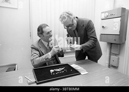 Commercianti di diamanti o valutatori?., Whizgle News, Dutch Desk, Paesi Bassi, 1950 - 2000. Questi argomenti sono mostrati nell'immagine. In un ambiente interno soffocato, due uomini sono impegnati in una discussione mirata a un tavolo di legno. Un uomo, seduto, è vestito con un abito grigio chiaro con una camicia con colletto, tenendo attivamente un oggetto e esaminandolo da vicino. La sua espressione trasmette curiosità e concentrazione. L'altro uomo sta accanto a lui, indossando un abito più scuro, piegando leggermente la testa verso l'individuo seduto. Sembra che stia usando un piccolo strumento portatile, forse una lente d'ingrandimento, per ispezionarne un altro Foto Stock