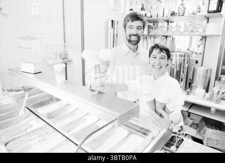 Gelateria Kors, pista di pattinaggio su ghiaccio Driehuis, Whizgle News, Dutch Desk, Paesi Bassi, 1950 - 2000 on 26-05-1989. Questi sono gli argomenti nell'immagine. In un'affascinante gelateria, due persone stanno dietro un bancone luminoso, entrambi macinati ampiamente. Contengono coni di gelato, ognuno dei quali ricoperto da un generoso turbinio di soffice panna montata bianca. L'uomo, con una barba leggera e capelli ben rifiniti, è vestito con una camicia button-up bianca e nitida. Il suo sorriso trasuda calore e cordialità. Accanto a lui, la donna sfoggia un semplice grembiule bianco su una camicia con colletto, i suoi capelli corti pervadono Foto Stock