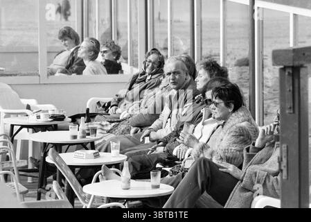 Seduti sulla terrazza al sole Zandvoort Terrace Zandvoort, Whizgle News, Dutch Desk, Paesi Bassi, 1950 - 2000 il 15-03-1986. Questi sono gli elementi dell'immagine. La scena raffigura un gruppo di persone sedute in una zona illuminata dal sole, probabilmente una passeggiata sul mare o una caffetteria. L'atmosfera è rilassata, con un senso di nostalgia evocato dall'abbigliamento vintage del popolo. Gli individui, principalmente gli adulti più anziani, sono vestiti con abiti a strati che includono maglioni e cappotti, suggerendo un ambiente ventilato. Si siedono a tavoli bianchi e rotondi, sui quali sono collocate delle bevande, possibilmente bicchieri di soda Foto Stock