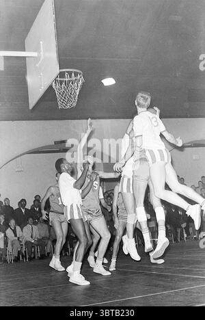 Partita di basket, Whizgle News, Dutch Desk, Paesi Bassi, 1950 - 2000 su 11-1967. Questi argomenti sono mostrati nell'immagine. La scena cattura un momento dinamico in una partita di basket, mostrando l'energia e l'intensità di uno sport competitivo. In primo piano, i giocatori in uniforme atletica sono impegnati in un gioco feroce, con un giocatore che salta in alto per fare un tiro verso il cerchio. Questo atleta si distingue in modo evidente contro gli altri, indossando una maglia di colore chiaro con il numero "13" sul retro, che contrasta con le tonalità più scure degli abiti dei suoi compagni di squadra. Gli altri giocatori sono posizionati a Foto Stock