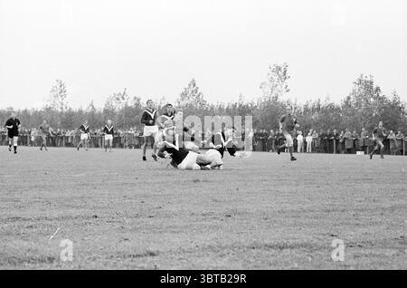 Partita di calcio., Whizgle News, Dutch Desk, Paesi Bassi, 1950 - 2000. L'immagine mostra questi argomenti. La scena cattura un momento intenso durante una partita di rugby ambientata su un campo erboso, circondato da una linea di alberi sparsa che crea un senso di apertura. I giocatori sono vestiti con uniformi contrastanti, alcune con maglie scure e altre con tonalità più chiare, a indicare le squadre in competizione. In primo piano, due giocatori sono impegnati in un placcaggio, uno in ginocchio, mentre l'altro sta saltando su di lui, mostrando la fisicità dello sport. Sudore e determinazione sono evidenti sui loro volti, aumentando il Foto Stock